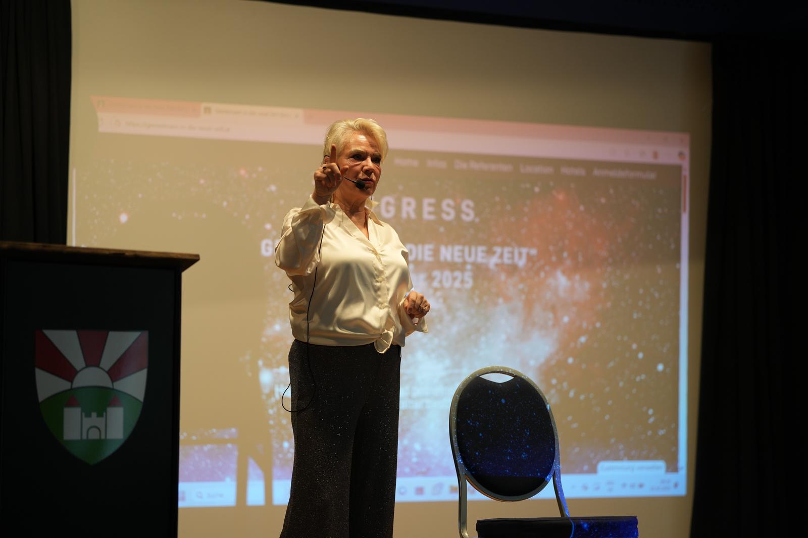 Vortragende steht auf der Bühne und trägt ein Thema vor. Im Hintergrund sieht man eine Präsentation auf der Leinwand und einen Stuhl