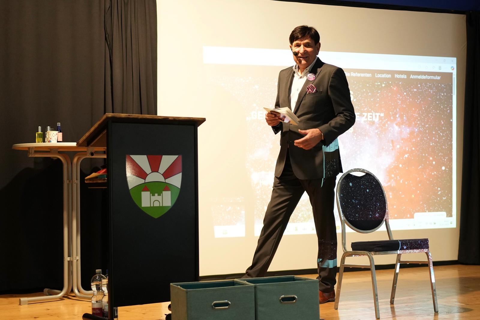 Veranstalter steht auf der Bühne und moderiert den Kongress. Hinter ihm ist eine Präsentation auf der Leinwand zu sehen