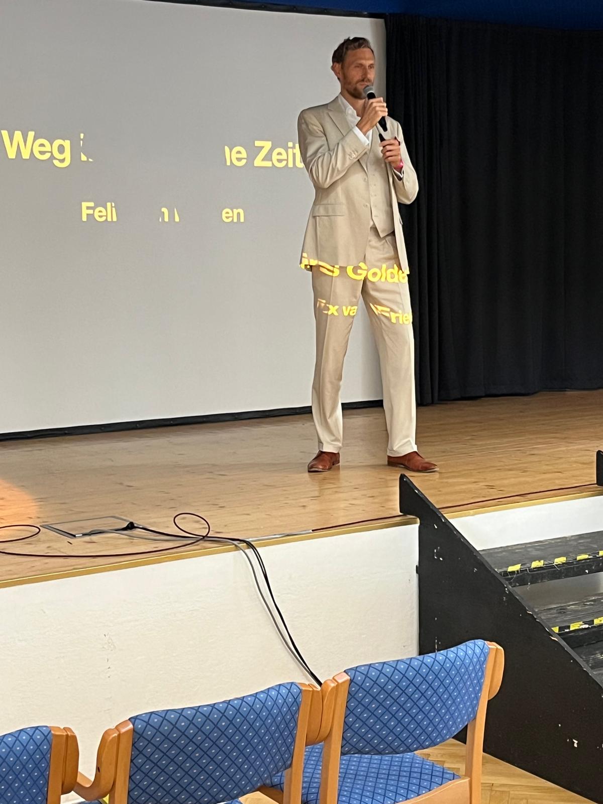 Vortragender Herr steht auf der Bühne, hinter ihm eine Leinwand mit Wörtern, vor der Bühne Zuschauerstühle
