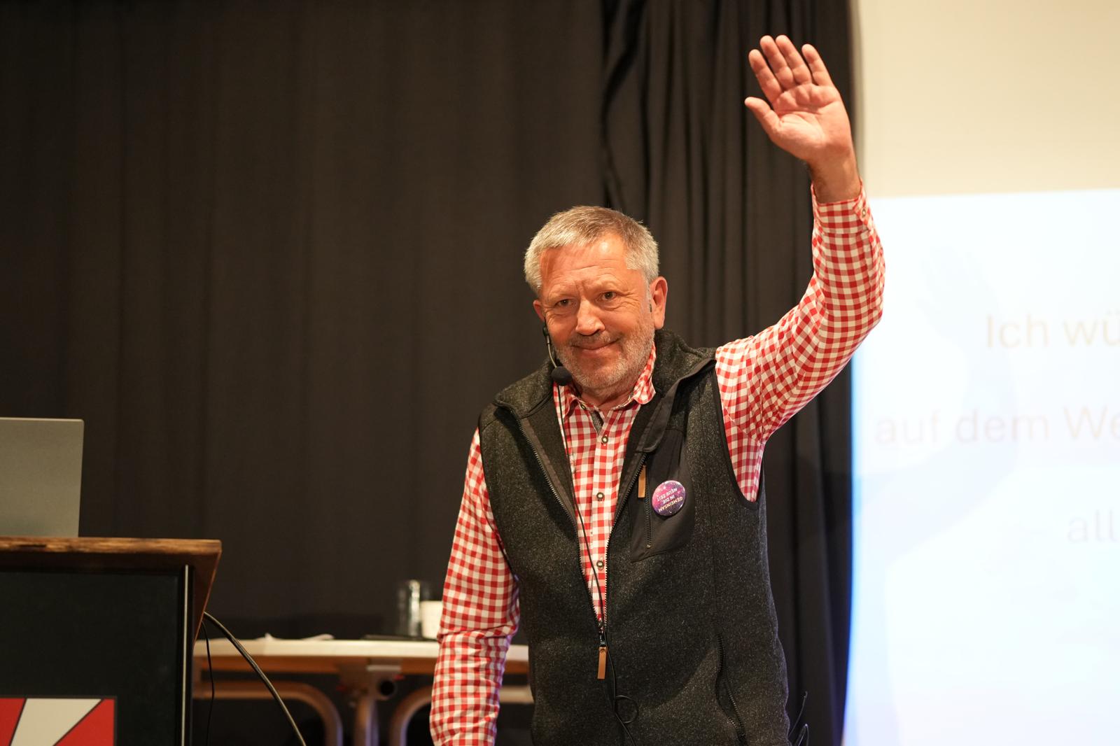 Vortragender grüßt das Publikum des Kongresses, trägt ein Karohemd und ein schwarzes Gilet, Präsentation im Hintergrund